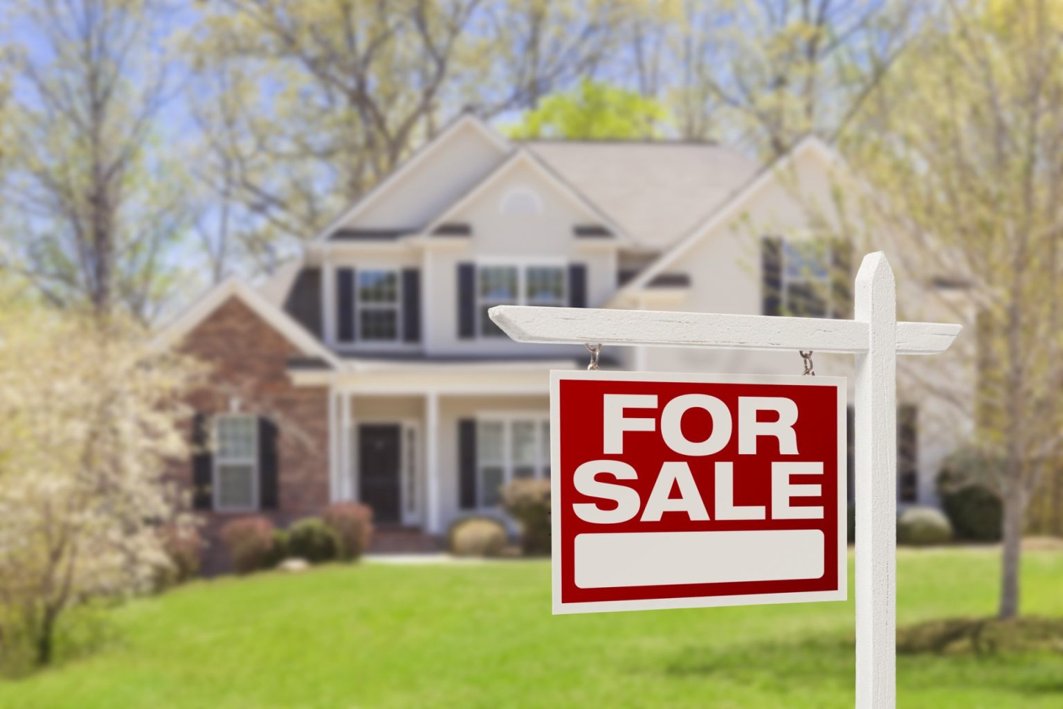 A close-up of a "For Sale" sign in front of a two-story home and green yard